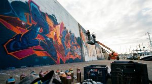 Fresque de graffiti colorée en cours de réalisation sur un mur de quai portuaire, bateaux de pêche en arrière-plan