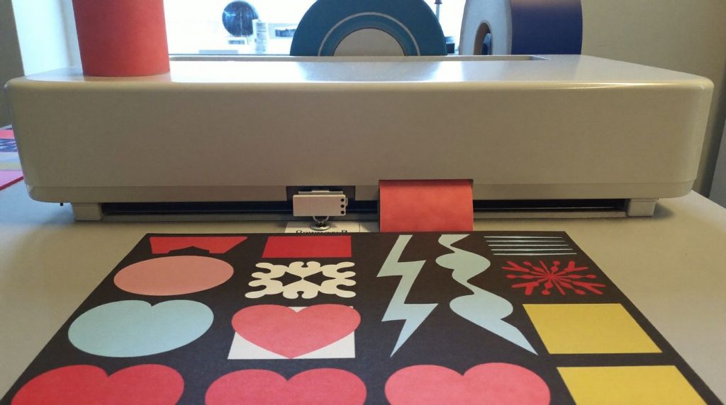 Plotter de découpe sur un établi, une planche de stickers aux formes colorées fraîchement découpées au premier plan